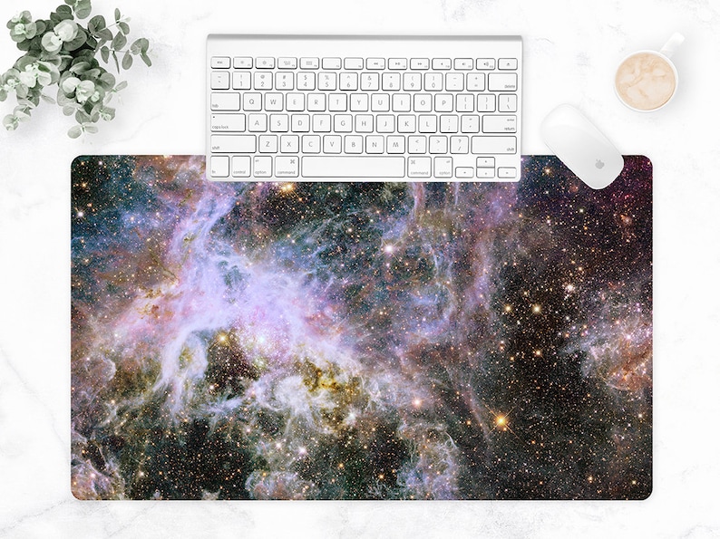 Puede incluir: Alfombrilla de rat&oacute;n rectangular con una imagen vibrante de una nebulosa con tonos morados, azules y dorados. Un teclado y un rat&oacute;n blancos est&aacute;n colocados encima de la alfombrilla. Tambi&eacute;n se ven una taza de caf&eacute; y una planta.