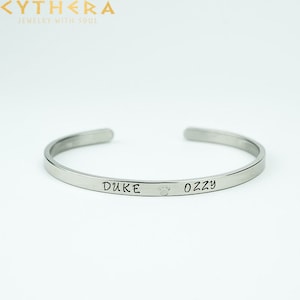 Könnte beinhalten: Silbernes Armreif-Armband mit den eingravierten Namen "Duke" und "Oz", getrennt durch einen Pfotenabdruck.