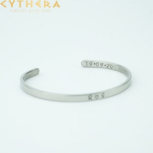 Può includere: Un bracciale a polsino color argento con la data incisa "18.08.20" e le iniziali "R ♡ S". Il gioiello è realizzato in metallo lucido e viene esposto su sfondo bianco. Il bracciale ha un design semplice ed elegante.