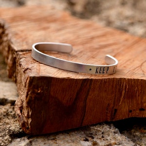 Pode incluir: Pulseira de bracelete de prata com as palavras "KEEP IT" gravadas na banda.