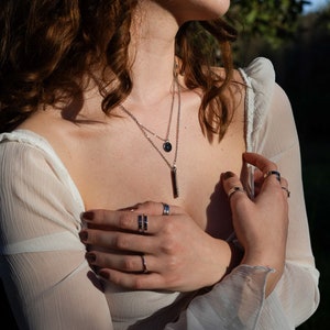 Op de afbeelding: Een vrouw draagt een wit doorschijnend topje met een gelaagde zilveren ketting en meerdere zilveren ringen aan haar vingers.