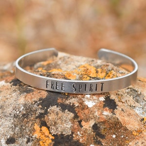 Pode incluir: Uma pulseira de metal prateada com as palavras "FREE SPIRIT" gravadas nela. A pulseira está apoiada em uma rocha com uma superfície marrom e amarela.