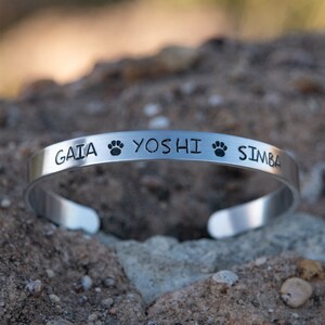 Pode incluir: Pulseira de prata com os nomes "GAIA", "YOSHI" e "SIMBA" gravados, separados por pegadas de patas.