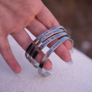 Könnte beinhalten: Eine Sammlung silberfarbener Metallarmreifen. Jeder Armreif trägt eine inspirierende Botschaft: "BE THE LIGHT", "THE C AND BACK", "TRUST IN LIFE" und "BE HAPPY". Die Armreifen sind gestapelt, was ihr schlichtes, elegantes Design hervorhebt.