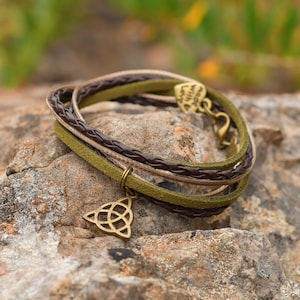 May include: A multi-strand bracelet with brown, olive green, and tan leather cords. The bracelet features a gold-coloured Celtic knot charm and a heart-shaped clasp. The bracelet is displayed on a textured rock surface.
