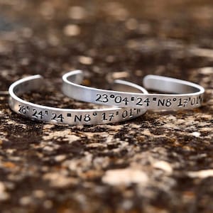 Puede incluir: Dos pulseras de plata con coordenadas grabadas. La pulsera superior dice "23°04' 24"N8°17'01'" y la pulsera inferior dice "28° 24' 24" N8° 17° 01"W".