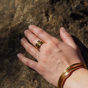 Pode incluir: Um anel de ouro com duas bandas, uma com um design de coração e a outra com uma inscrição de palavras. O anel é usado no dedo de uma pessoa. A pessoa está usando uma pulseira de ouro no pulso.