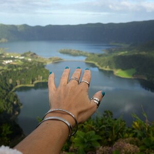 May include: A hand with teal nail polish reaches out towards a scenic view of a lake and green hills. The hand is wearing several silver rings and a silver bracelet.