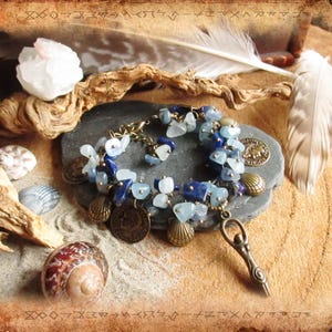 Può includere: Un braccialetto blu e argento con un ciondolo di una donna in piedi su una spirale. Il braccialetto è realizzato con perline di acquamarina, lapislazzuli e conchiglie. Il braccialetto è su una superficie di legno con una piuma e conchiglie.