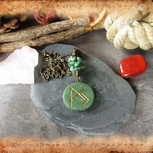 May include: A green stone pendant with a gold chain and a gold rune symbol. The pendant is surrounded by green stones and is on a grey stone surface.