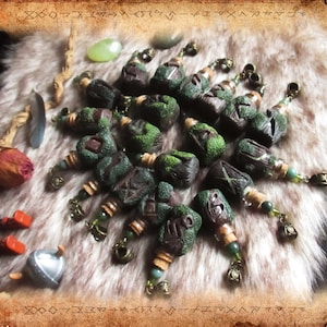 Könnte beinhalten: Eine Sammlung von 16 kleinen, grünen, braunen und goldenen Charms mit in sie geschnitzten Runen. Die Charms sind auf einer weißen, flauschigen Oberfläche angeordnet.