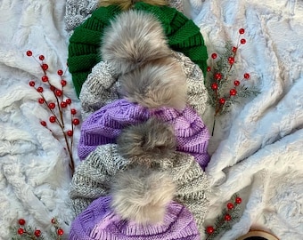 May include: Five knitted hats with faux fur pom poms in various colors, including purple, gray, and green. The hats are stacked on top of each other on a white fluffy surface.