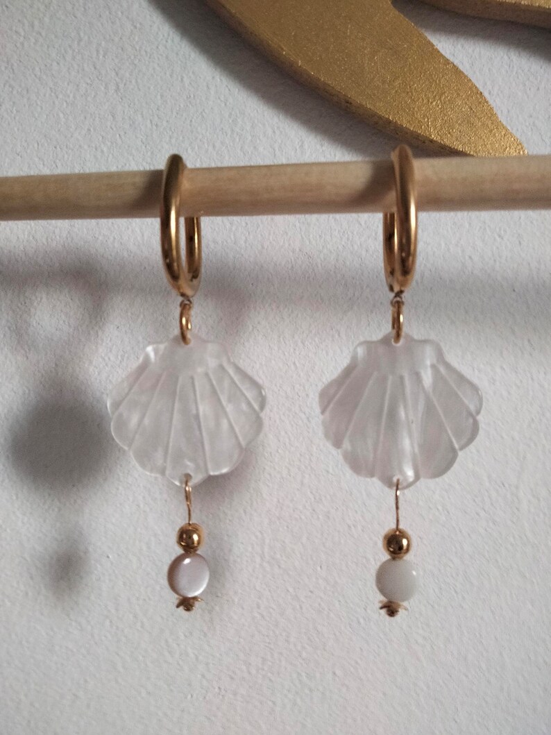 May include: A pair of gold hoop earrings with a white seashell charm and a white pearl bead dangling from each hoop.