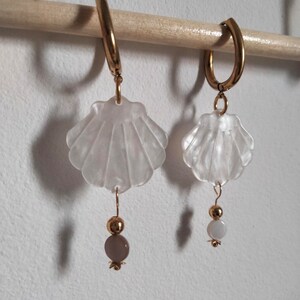 May include: A pair of gold hoop earrings with white shell charms and a single bead dangling from each charm. The beads are different colors, one is white and the other is a light brown.