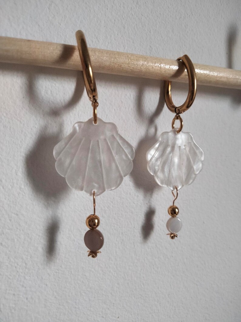 May include: A pair of gold hoop earrings with white shell charms and a single bead dangling from each charm. The beads are different colors, one is white and the other is a light brown.