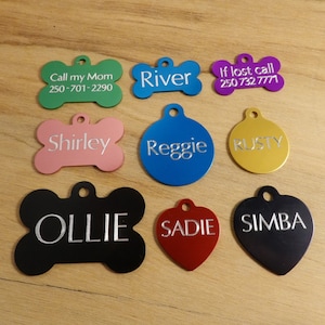 May include: A selection of personalised dog tags in a variety of shapes and colours, including bone, circle and heart shapes. The tags are engraved with names such as "River", "Reggie", "Rusty", "Ollie", "Sadie" and "Simba". Some tags also include phone numbers.