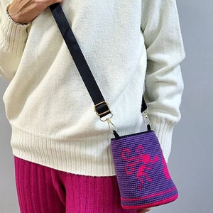 May include: A purple crocheted bag with a pink design hangs from a black strap on a person's shoulder. The bag is worn over a white sweater and pink pants.