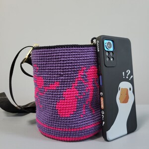 May include: A purple crocheted crossbody bag with a pink design and a black phone case with a cartoon goose and the text "!?!?" and "Goose" on the back.