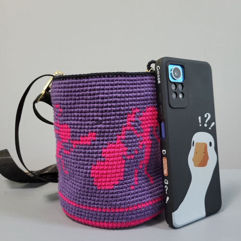 May include: A purple crocheted crossbody bag with a pink design and a black phone case with a cartoon goose and the text "!?!?" and "Goose" on the back.