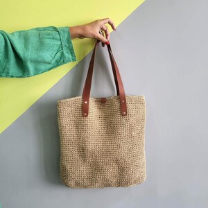 May include: A rectangular tote bag crafted from a natural tan woven fiber material. The bag features two long, smooth brown leather shoulder straps securely attached to the top edge with silver-toned metal rivets on each side of the bag.