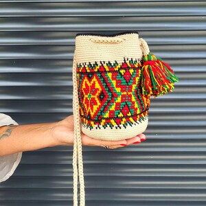 May include: A cylindrical crochet bucket bag in a light beige color featuring a vibrant geometric pattern in red, yellow, and green. The bag includes a matching beige drawstring closure and a large, multi-colored tassel in red, yellow, and green.