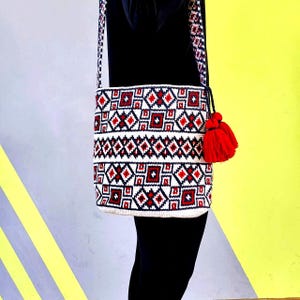 May include: A white, red, and navy blue geometric pattern crossbody bag with a red tassel. The bag has a woven design with a long strap. The bag is hanging against a yellow and gray background.