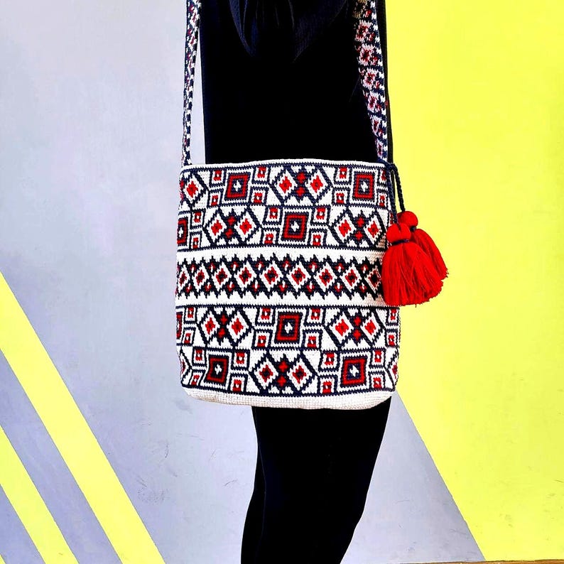May include: A white, red, and navy blue geometric pattern crossbody bag with a red tassel. The bag has a woven design with a long strap. The bag is hanging against a yellow and gray background.