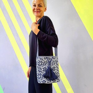 May include: A navy blue and white geometric patterned shoulder bag with a navy blue tassel. The bag has a long strap and is held by a person. The background is a mix of gray, yellow, and white.
