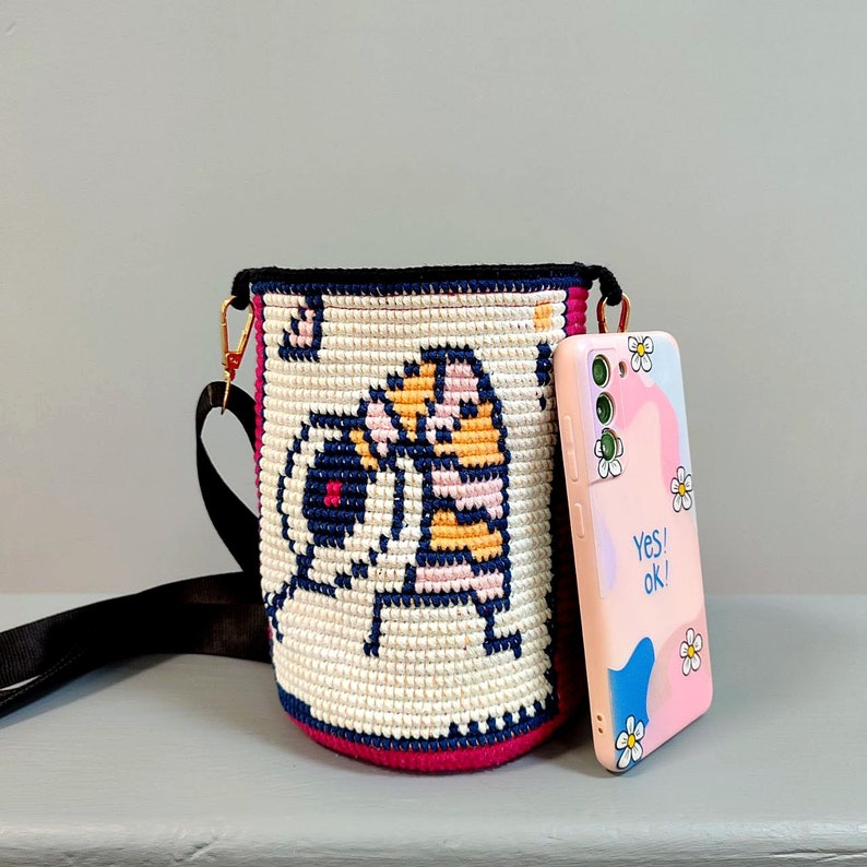 May include: A white and black crocheted bucket bag with a cartoon character design. The bag has a black strap and is paired with a pink and white phone case with the text "Yes! Ok!"