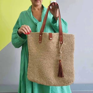 May include: A rectangular tote bag made of tan woven material featuring two brown leather shoulder straps attached with silver-toned metal rivets. A central leather tab with a snap closure secures the top, and a brown leather tassel hangs from one strap.