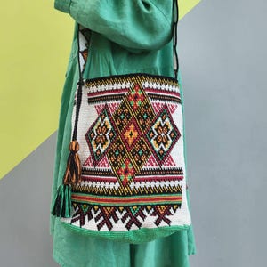 May include: A crochet shoulder bag featuring a cream-colored base with intricate geometric patterns in black, red, yellow, and green. The bag includes a long strap and a decorative tassel with black and orange threads hanging from the side attachment.