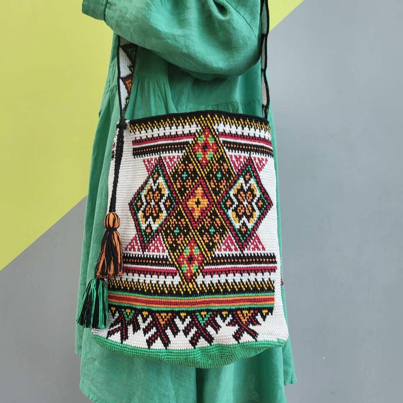 May include: A crochet shoulder bag featuring a cream-colored base with intricate geometric patterns in black, red, yellow, and green. The bag includes a long strap and a decorative tassel with black and orange threads hanging from the side attachment.