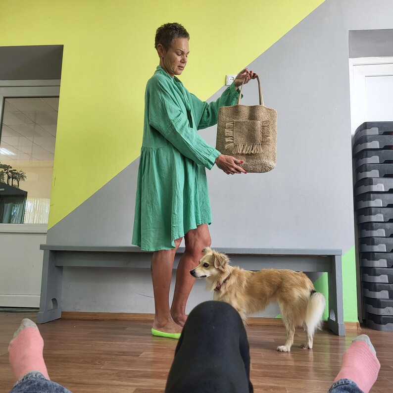 May include: A light green long-sleeved dress with a loose fit and a hemline that sits above the knee. The dress is paired with a rectangular beige woven tote bag featuring a front pocket with frayed edges and two thin shoulder straps.