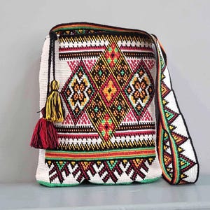 May include: A crochet shoulder bag featuring a white base with intricate geometric patterns in black, red, yellow, and green. The bag includes a long matching shoulder strap and a decorative tassel detail in yellow and red hanging from the side.