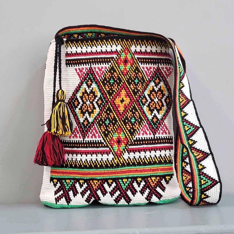 May include: A crochet shoulder bag featuring a white base with intricate geometric patterns in black, red, yellow, and green. The bag includes a long matching shoulder strap and a decorative tassel detail in yellow and red hanging from the side.