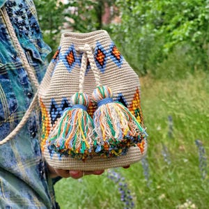 May include: A beige crochet bucket bag featuring a geometric pattern in orange, blue, and yellow. The bag includes a drawstring closure with two thick braided cords, each ending in a large, multi-colored tassel made of loose, vibrant threads.