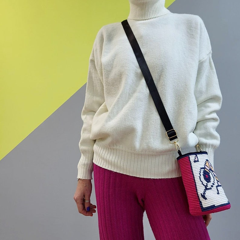 May include: A white turtleneck sweater with a black shoulder strap and a small pink and white crocheted bag with a colorful design. The bag has a gold clasp and a black strap.