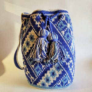 May include: A blue and white crochet bucket bag with a geometric pattern. The bag features a drawstring closure and two decorative tassels. The strap is approximately 30 inches long. The bag is a vibrant accessory.