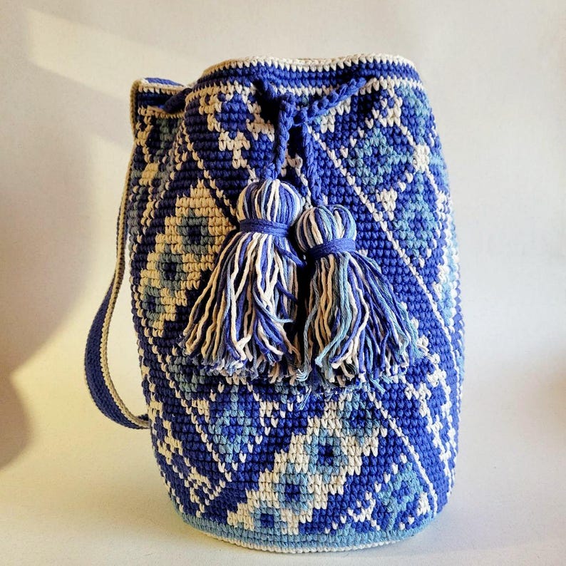 May include: A blue and white crochet bucket bag with a geometric pattern. The bag features a drawstring closure and two decorative tassels. The strap is approximately 30 inches long. The bag is a vibrant accessory.