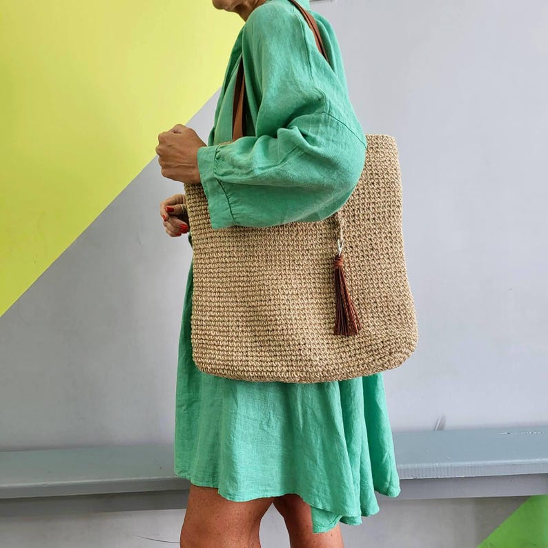 May include: A bright green long-sleeved dress with a loose fit and a large, rectangular, tan woven tote bag featuring a brown leather strap and a matching brown tassel hanging from the side of the bag, worn over the shoulder.