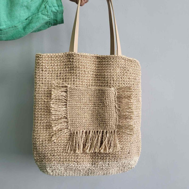 Large jute tote bag held by a woman, showing the handmade crochet texture, front pocket and long shoulder straps.