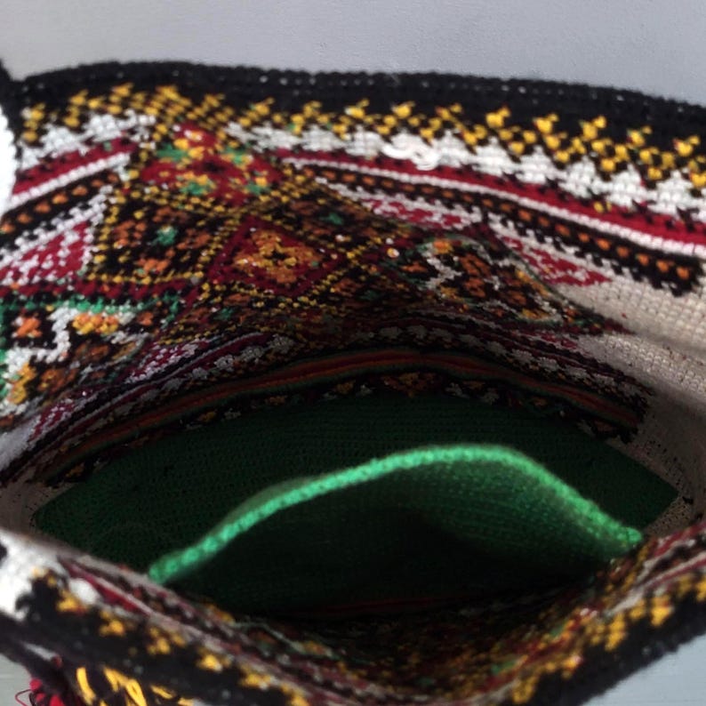 May include: A textile bag featuring a black, white, red, and yellow geometric embroidered pattern on the exterior. The interior of the bag is lined with a solid green fabric, and the top edge is finished with a black crocheted border.