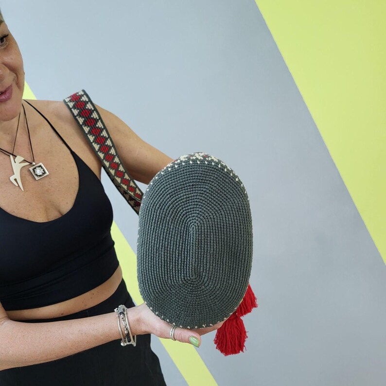 May include: A gray crocheted bag with red tassels. The bag has a round bottom and a woven design.