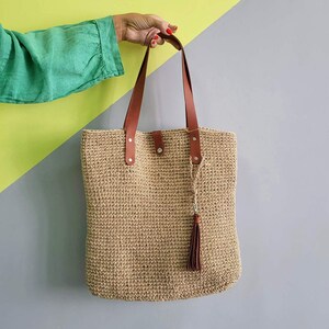 May include: A rectangular tote bag made of woven tan natural fiber material. The bag features two long brown leather shoulder straps attached with silver metal rivets and a matching brown leather tassel hanging from one of the strap attachment points.