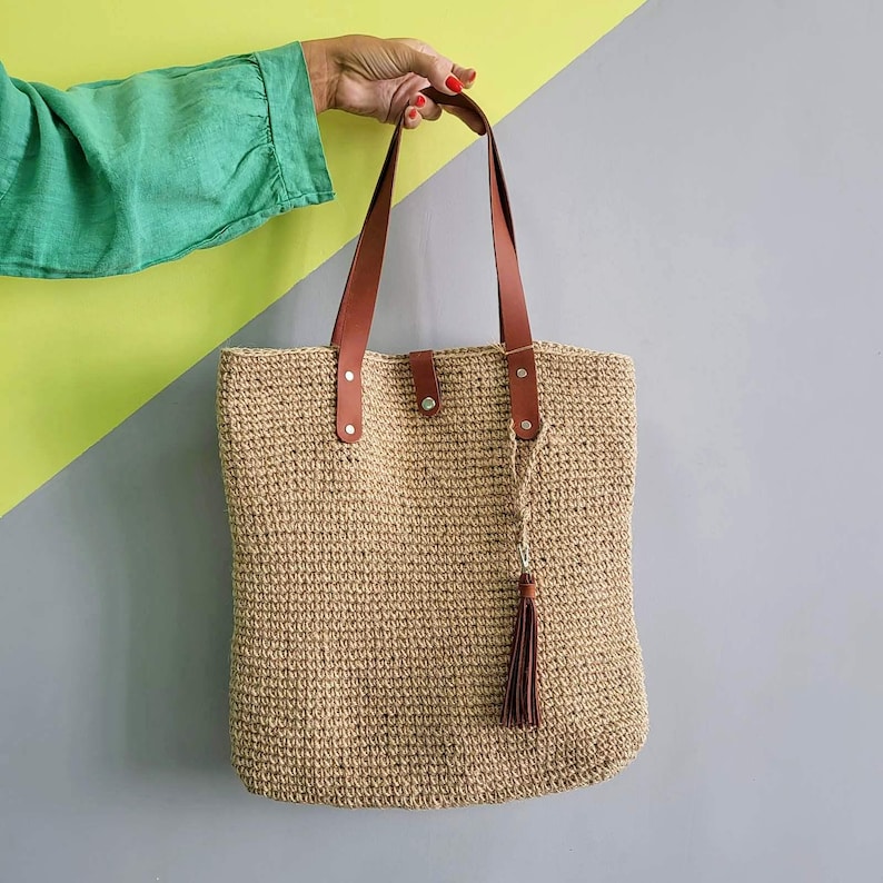 May include: A rectangular tote bag made of woven tan natural fiber material. The bag features two long brown leather shoulder straps attached with silver metal rivets and a matching brown leather tassel hanging from one of the strap attachment points.