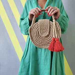 May include: A round, tan, woven jute handbag featuring a structured circular body, a top handle, and a flap closure. The bag is accented with two vibrant red tassels hanging from a braided cord attached to the side, creating a textured appearance.