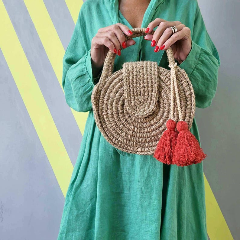 May include: A round, tan, woven jute handbag featuring a structured circular body, a top handle, and a flap closure. The bag is accented with two vibrant red tassels hanging from a braided cord attached to the side, creating a textured appearance.