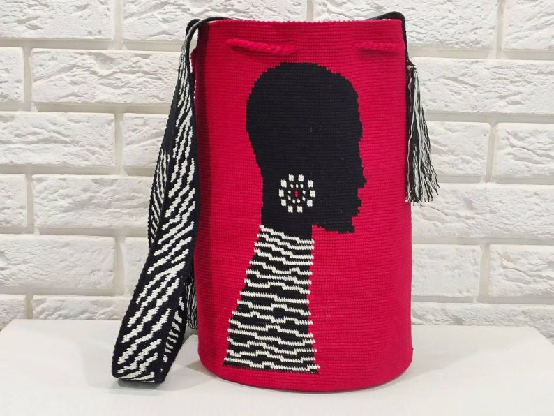 May include: A red crocheted bag with a black and white patterned strap. The bag features a silhouette of a person with a black and white patterned design and a red circle earring.