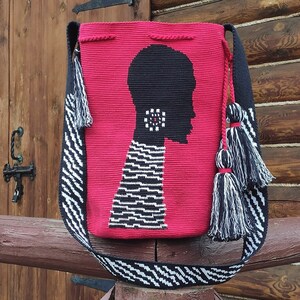 May include: A red crocheted bag with a black and white silhouette of a person's head. The bag has a black and white striped strap with tassels.