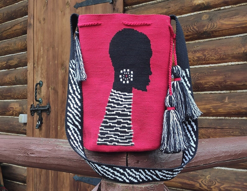 May include: A red crocheted bag with a black and white silhouette of a person's head. The bag has a black and white striped strap with tassels.